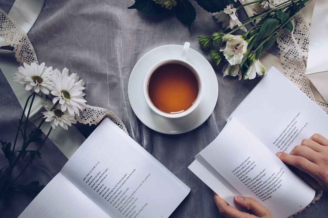 Hands holding an open book on a bed beside a cup of tea and white flowers